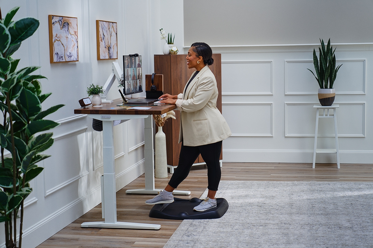 Person standing on an Active Anti-Fatigue Mat at an Uplift Standing Desk doing work and smiling.