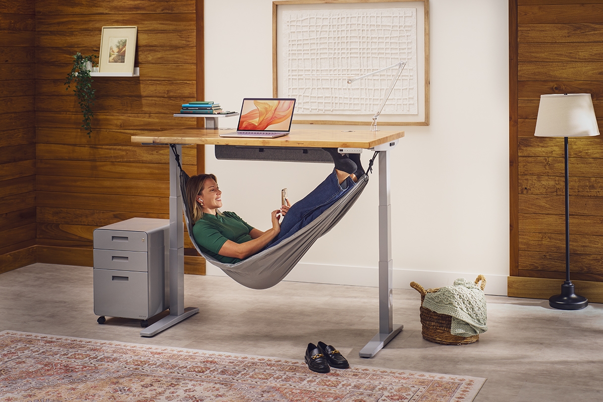 Person relaxing on their phone while laying in an Under Desk Hammock attached to an Uplift Standing Desk.