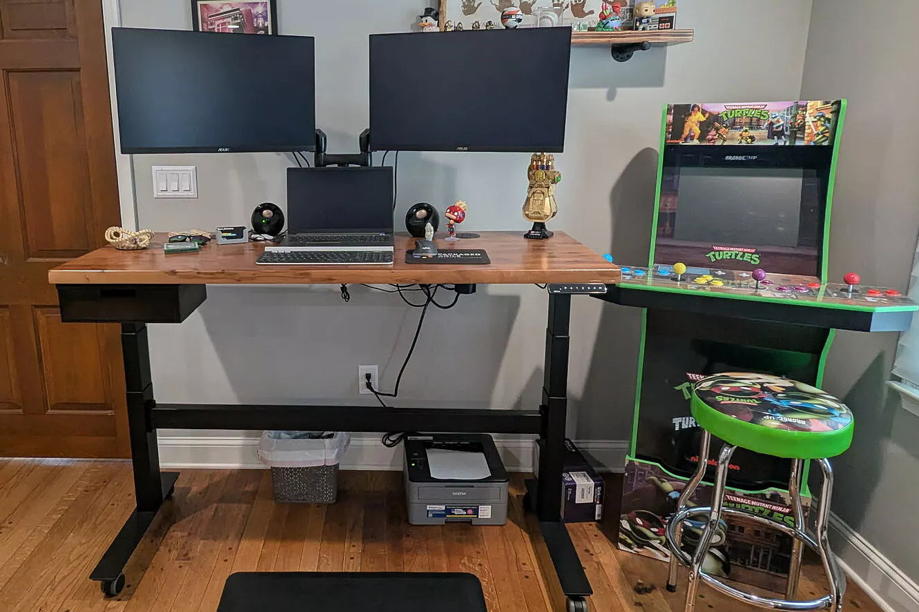 A home office featuring an Uplift Standing Desk holding two monitors and a laptop, next to a Teenage Mutant Ninja Turtles arcade machine.