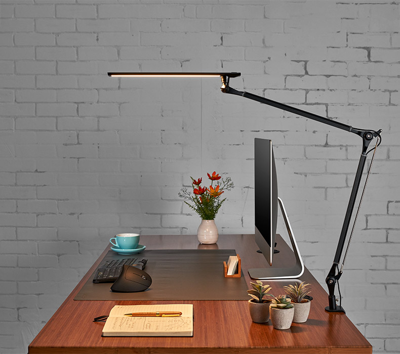 A black E7 Desk Lamp clamped to the back of a bamboo uplift standing desk