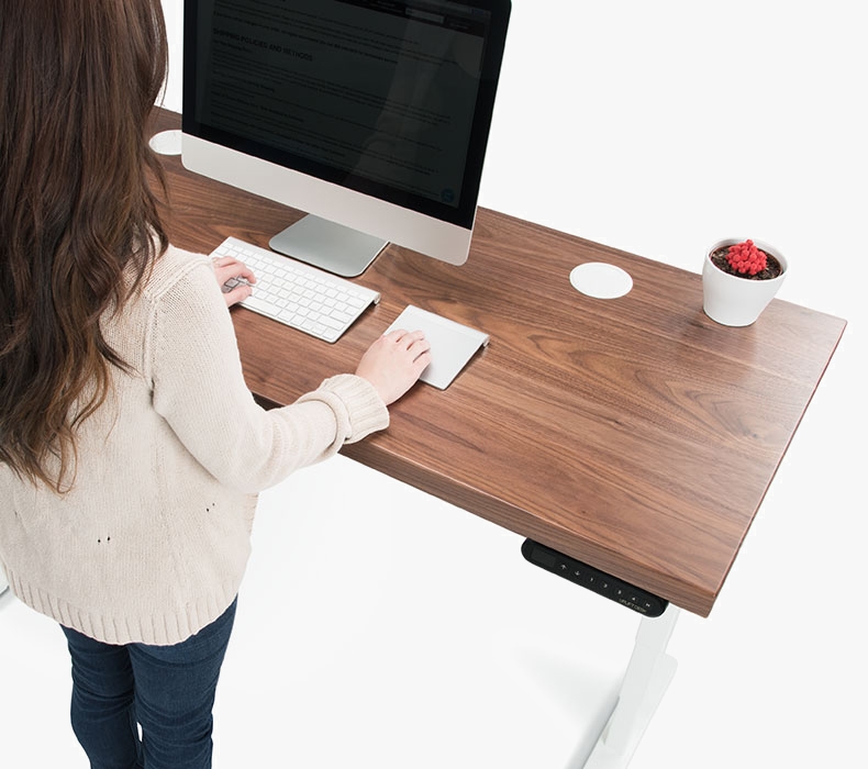 A woman on her computer stands at her UPLIFT Desk with a walnut desktop shown and white grommet covers.