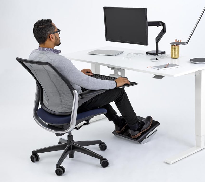 A man comfortably resting his feet on the Humanscale FR500 Ergonomic Foot Rocker Footrest in black finish while working.