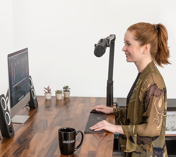 A person working at an UPLIFT Desk speaking into a microphone supported by a Clamp-On Microphone Arm