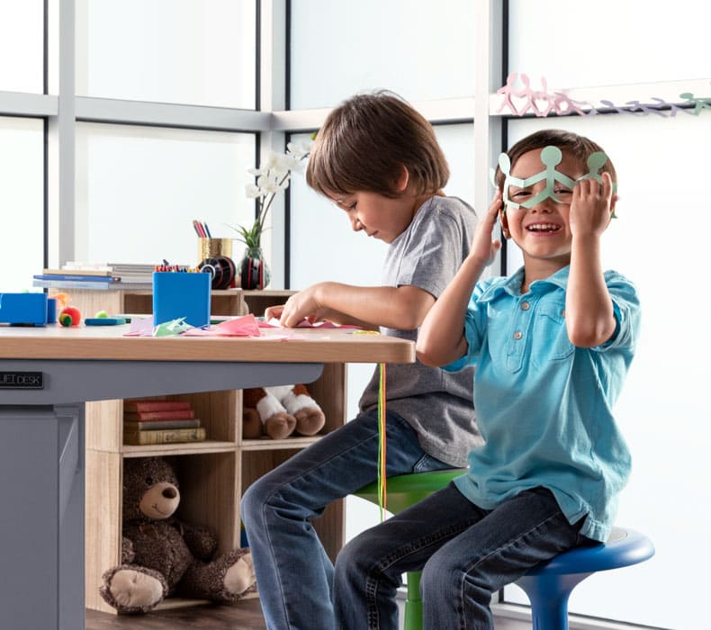 Two kids playing at a height adjustable desk; one is coloring, and one is holding up a paper mask